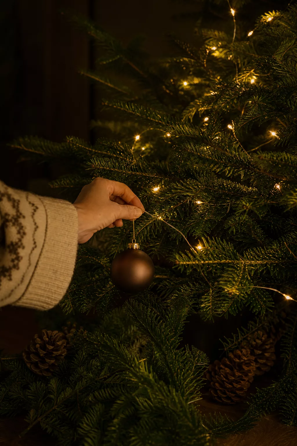 Hand hängt Christbaumkugel an beleuchteten Weihnachtsbaum abends auf