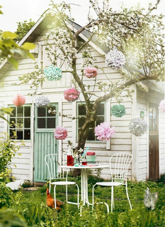 weißes-Gartenhäuschen-Gras-Tisch-Stühle-Baum-Papierblumen-Huhn