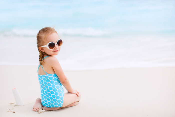 Little girl with bottle of sun cream sitting at tropical beach