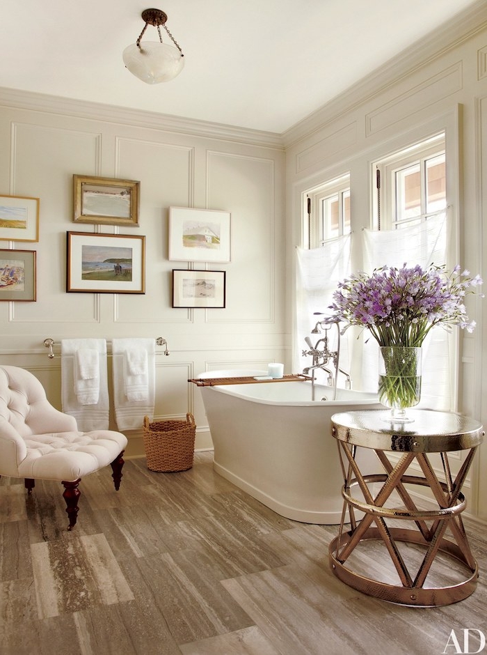 Luxuriöses Badezimmer, weiße Badewanne aus Keramik und metallener Tisch, weißer Ohrensessel und Rattan Korb auf dem Boden, Bilder an der Wand