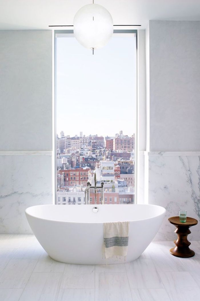 Bad modern gestalten, weiße Badewanne aus Keramik und kleiner Holztisch, großes Fenster und Marmorwand 