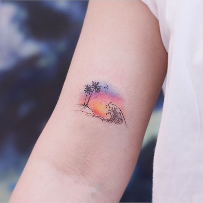 Kleines farbiges Tattoo am Oberarm, Welle und kleine Insel mit zwei Palmen bei Sonnenuntergang 