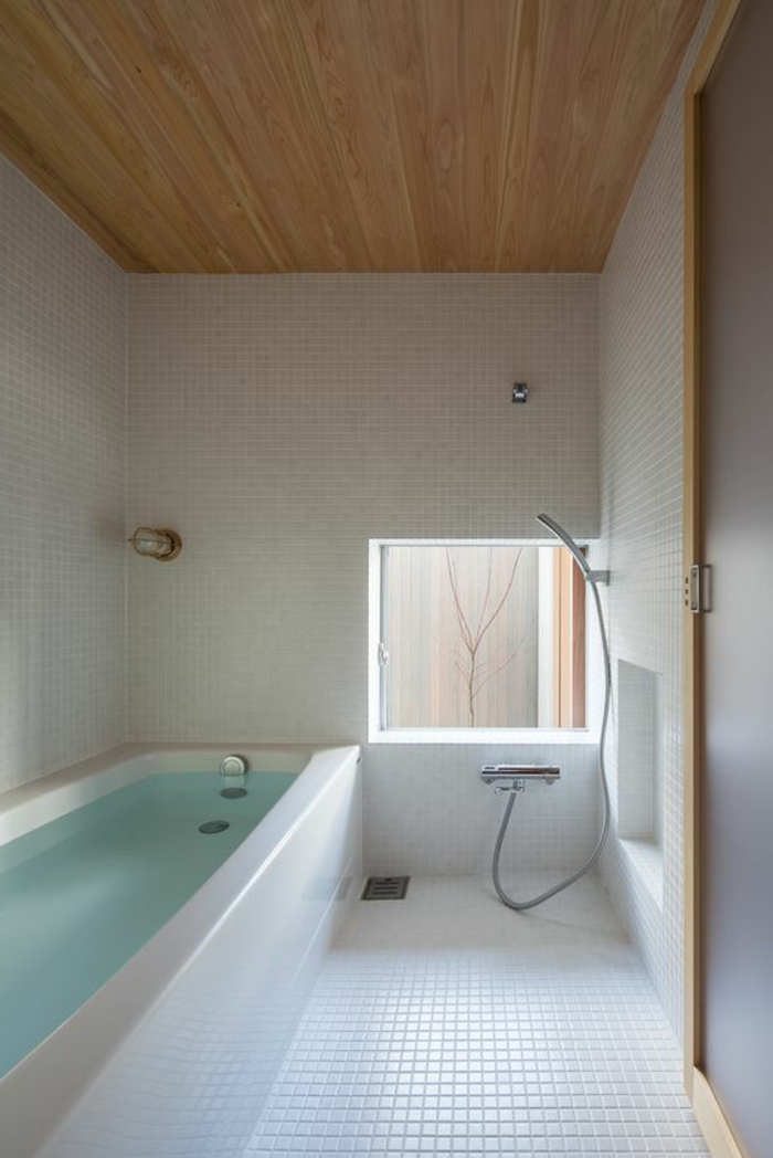 Badezimmer mit eingebauter Badewanne, weißen Fließen, viereckigem Fenster neben der Dusche, Holzdecke