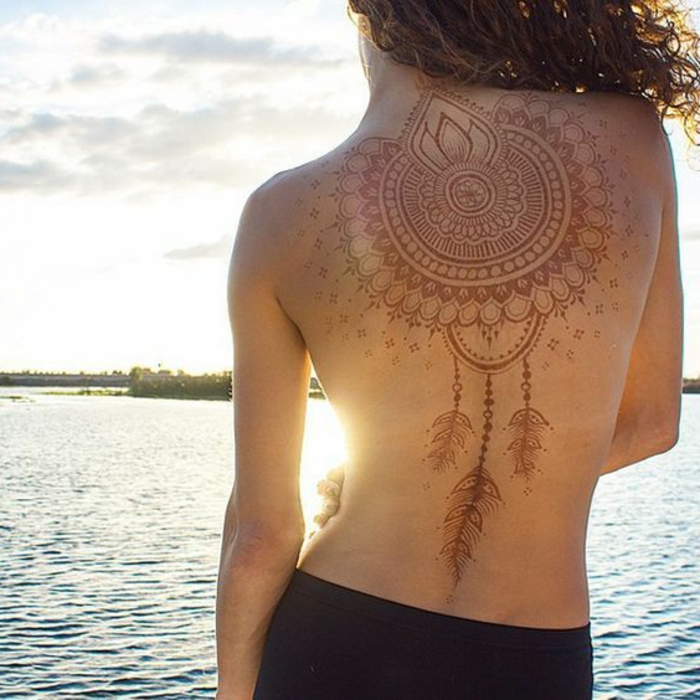 eine Frau am Strand mit temporärem Traumfänger Tattoo aus Henna in roter Farbe, Federn Tattoo, lange lockige Haare