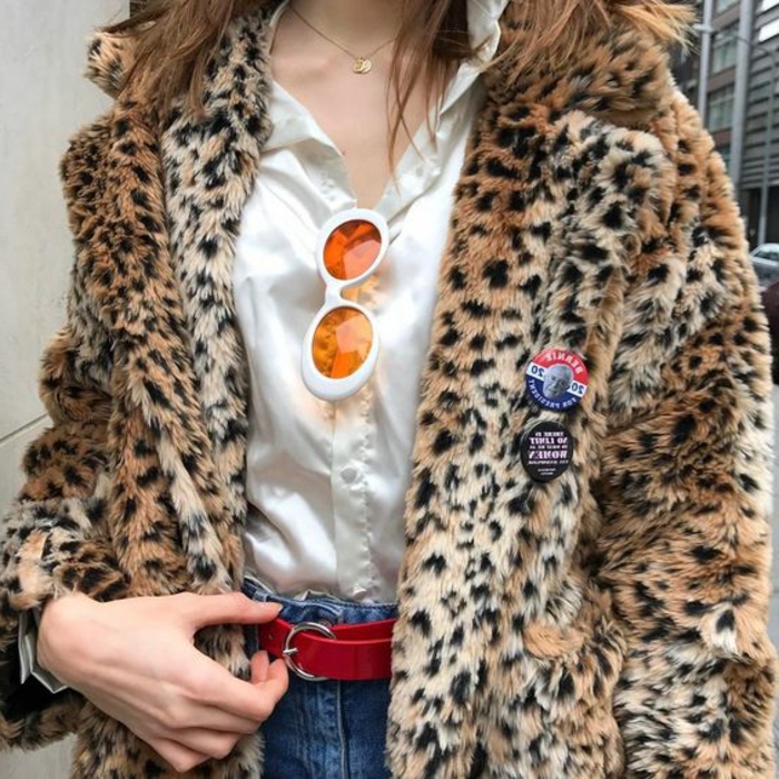 80er Frau mit einer Jacke mit Leopardprint, weißem Satinhemd, Jeans mit rotem Gürtel, weiße Sonnenbrille mit orangen Gläsern