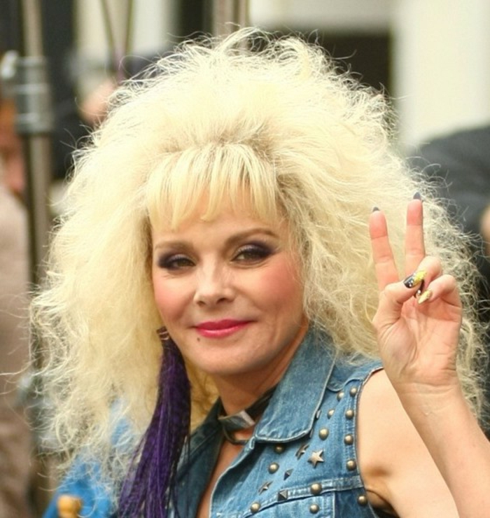 Kim Cattrall mit 80er Outfit: Denimjacke mit Metallkapseln und -sternen, topiertes blondes Haar mit Pony, riesige Ohrringe