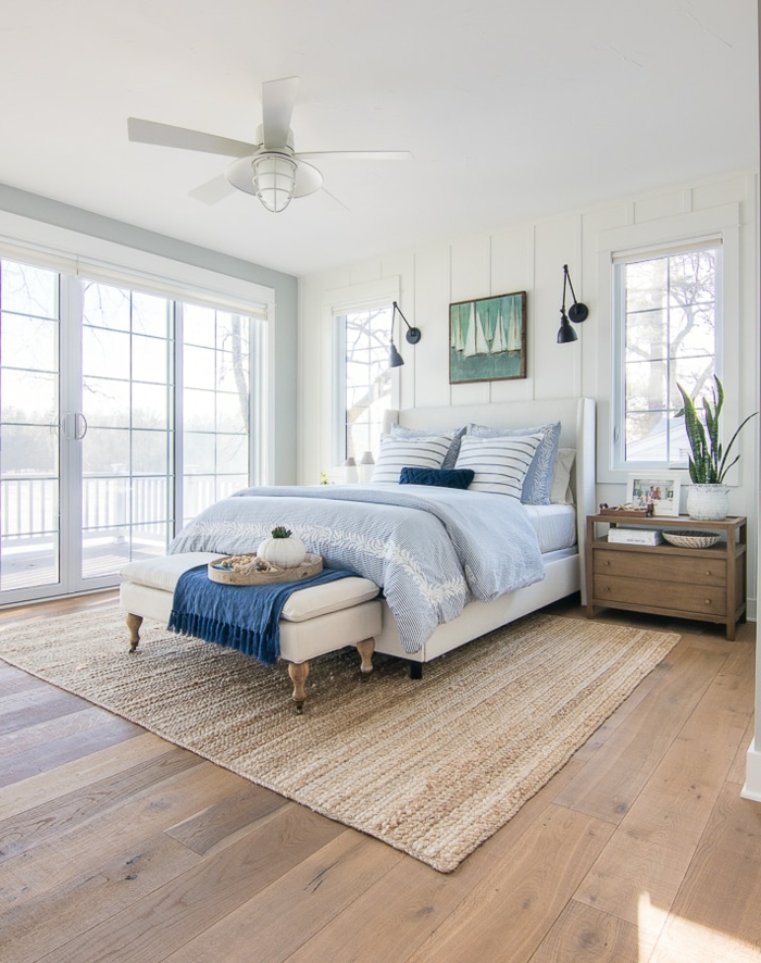 großes Schlafzimmer maritim, großes Bett mit blauen Bettwäschen, gemaltes Bild von einem Segelboot, hellbrauner Teppich, Kommode aus Holz