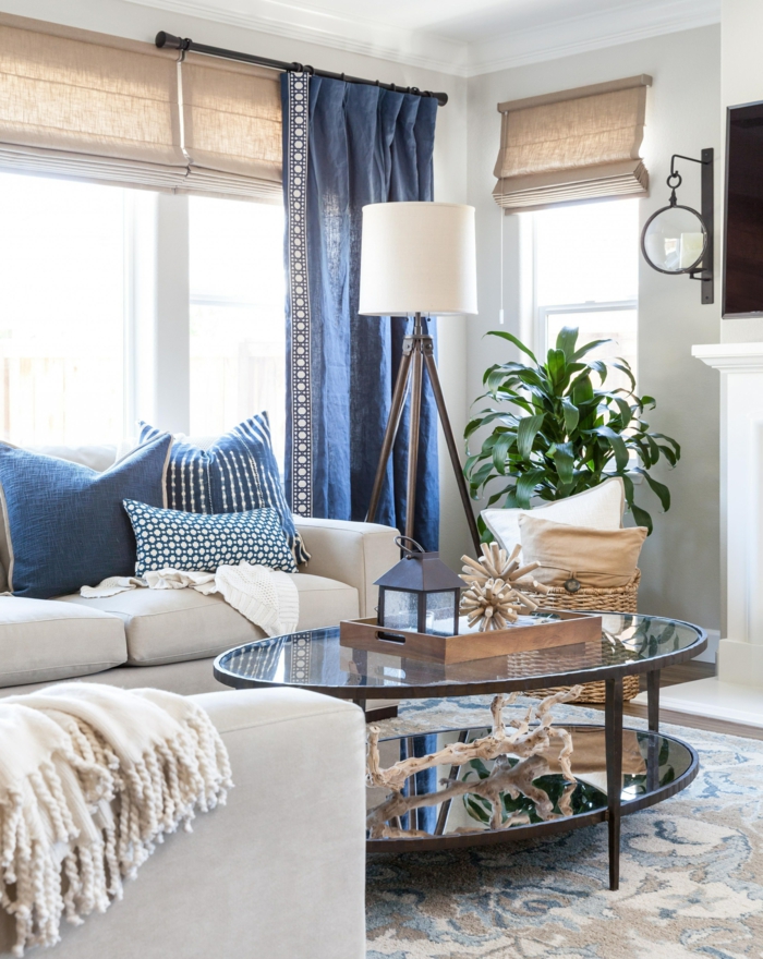 Wohnzimmer maritimer Stil, blaue Gardinen und Kissen, Sofa in beige, grüne Pflanze, runder Glastisch, Teppich mit blauen Motiven
