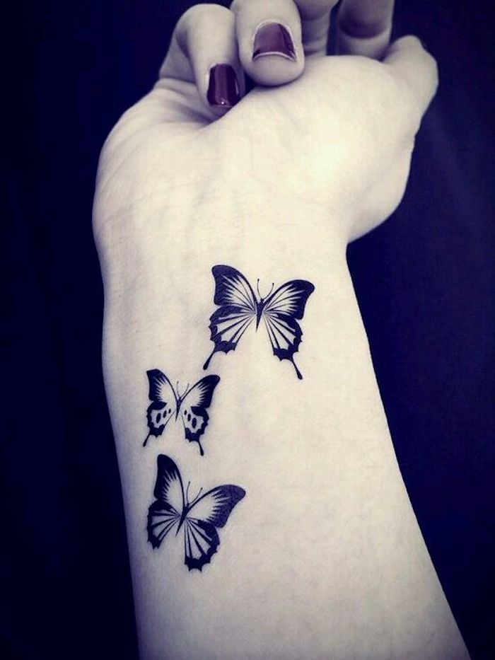 das ist eine der besten ideen zum thema schmetterling tattoo, die ihnen sehr gut gefallen kann - drei kleine, schwarze, fliegende schmetterlinge auf dem handgelenk einer hand einer jungen frau 