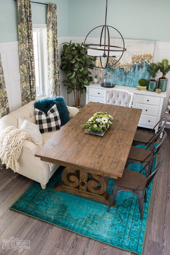gemütliches Wohnzimmer im Landhausstil, massiver Holztisch, verspielter Kronleuchter, blauer Teppich, grüne Zimmerpflanzen, weiße Schrank und Sofa