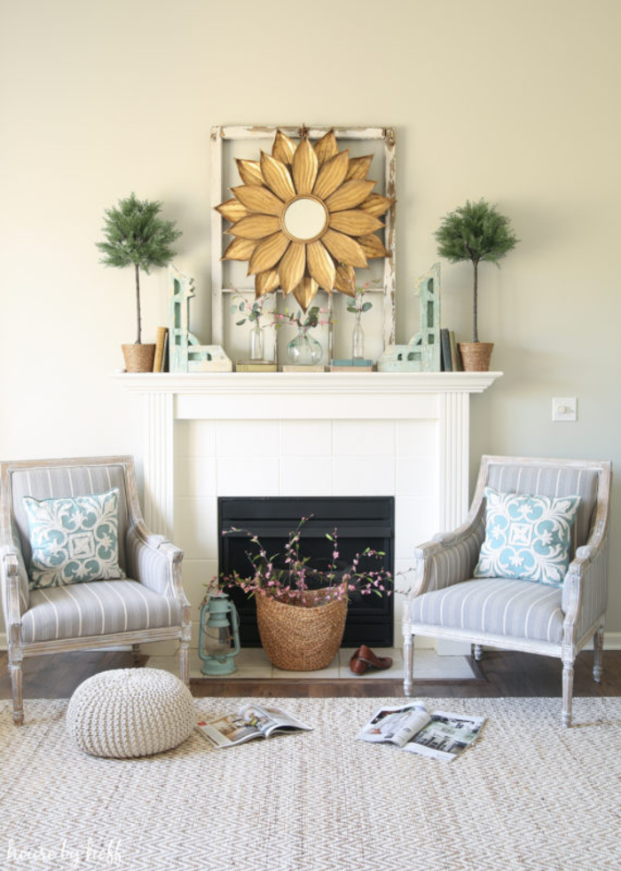 Wohnzimmer in zarten, hellen Farben, Kamin und zwei Sessel, Spiegel im Rahmen als Sonne, Rattankorb mit Blütenzweigen