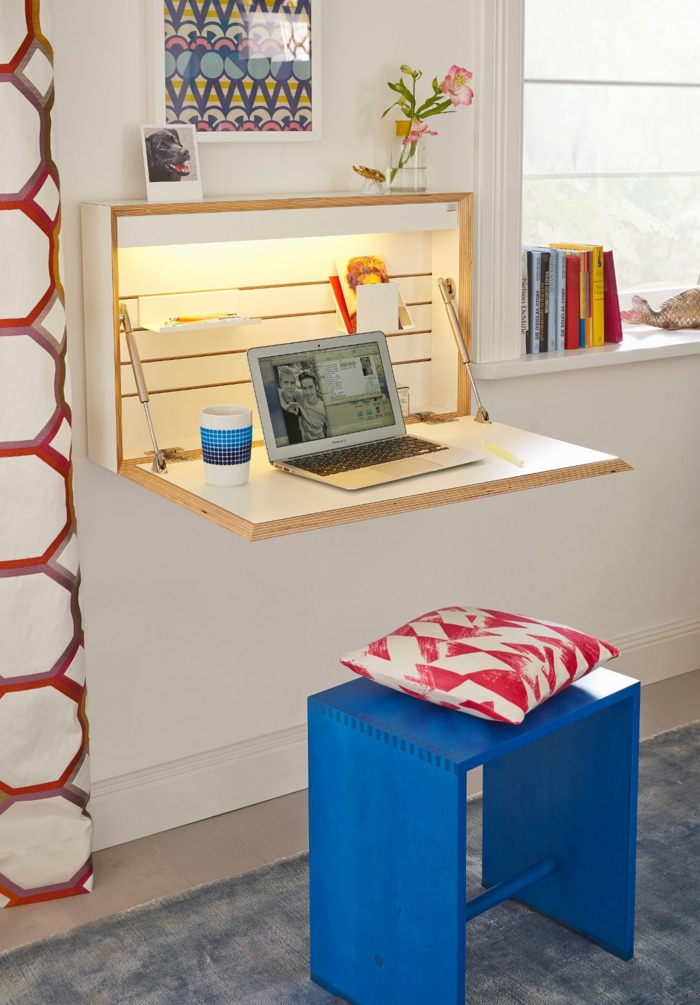 Schreibtisch für kleine Räume, funktionelle und praktische Ideen, blauer Holzhocker, weiß-rotes Kissen, Bücher am Fenster, Laptop und Porzellantasse auf dem Tisch