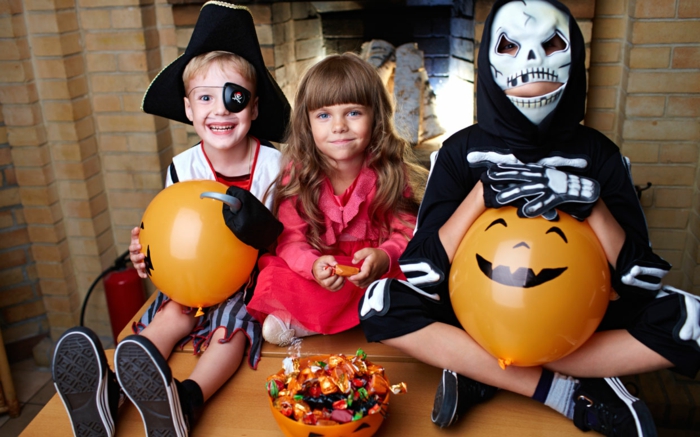 Drei süße Kinder mit Halloween Kostümen, orange Luftballons als Kürbisse, Schale, voll mit Süßigkeiten