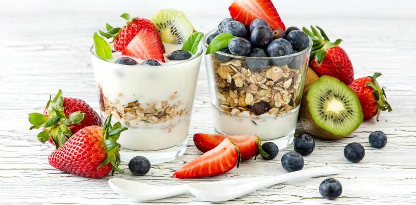 Joghurt im Glas mit drei Sorten von Früchten - Kiwi, Erdbeeren, Blaubeeren, serviert mit Haferflocken, weiß gestrichener Holztisch, zwei kleine Porzellanlöffel
