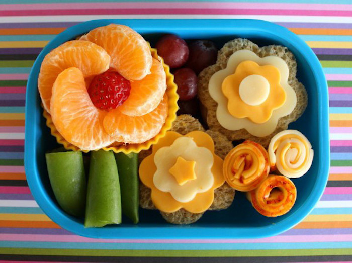 zwei Butterbrote in der Form von Blumen, gelbe und weiße Käse, frisches Obst, bunte Tischdecke mit Streifen in allen Farben