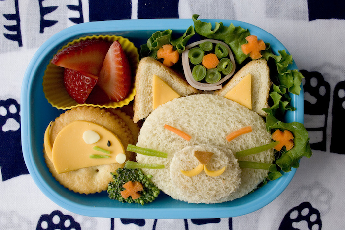 Mittagessen für kleine Kinder: Sandwich in der Form eines Katzengesichts, Schnurrhaare aus grüner Paprika, gewürfelte Erdbeeren, Kekse mit Käse, Tischdecke mit Katzenpfoten-Print