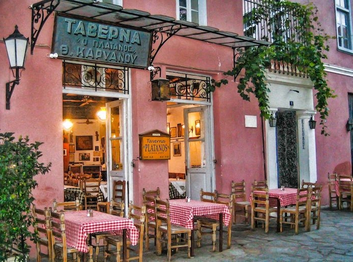 athen sehenswürdigkeiten taverna tawerna in athen gebäude in rosarot gemalen grüne pflanzen auf dem balkon über dem restaurant kleine tische mit kariierten tischdecken mediterranes essen probieren essen und genießen