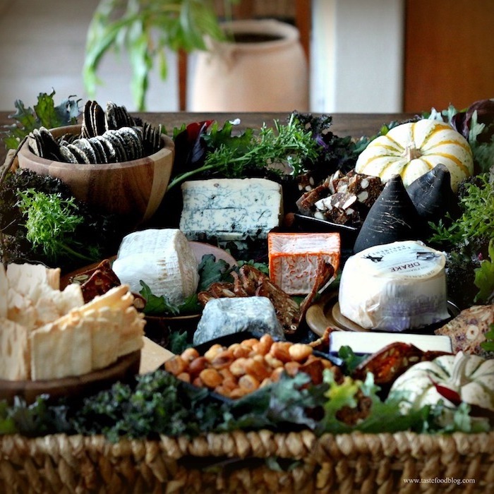 ein Holztisch voll von verschiedenen frischen Gewürzen, Käse in Papierverpackung, Käse in roter Wachsverpackung, Kekse mit Schokolade und Puderzucker, zwei weiße Kürbisse mit grünen und gelben Streifen auf der Kürbiskruste, Wand mit Holzverkleidung