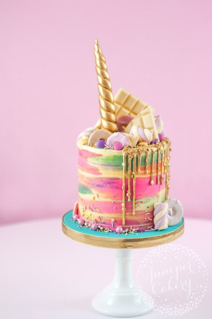 eine bunte regenbogenfarbene einhorn torte mit einer weißen schokolade und mit einem großen goldenen langen gelbenen horn und eine pinke wand