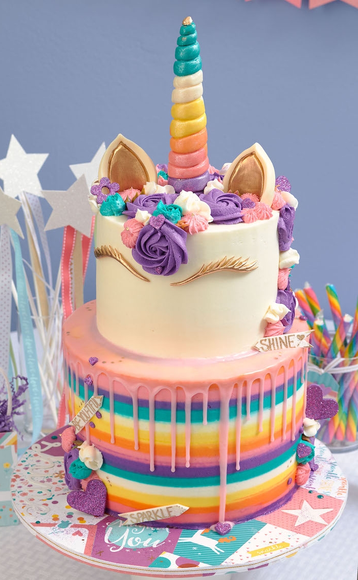 idee für eine zweistöckige einhorn torte mit einem weißen horn mit einem langen regenbogenfarbenen horn und mit einer mähne aus lila und kleinen blauen und pinken rosen - idee für einhorn kuchen deko