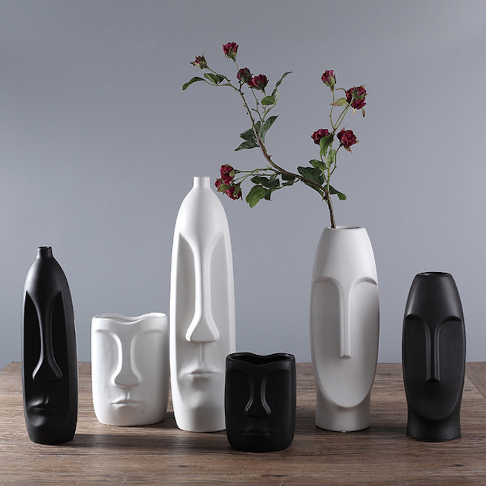 Geschenke zur Einweihung: sechs dekorative Vasen mit skandinavischem Design, die Gesichtsskulpturen ähneln, drei weiße und drei schwarze Blumenvasen in verschiedenen Größen, Holztisch mit Lacküberzug, hellgraue Wand als Hintergrund, rote Nelken in einer Blumenvase