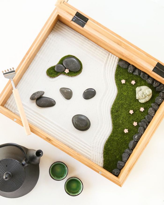 ein Holzkasten mit weißem Sand und kleinen grauen gerundeten Flußsteinen, eine Minifigur von Buddha aus Nephritis, kleiner Zengarten in einer hölzernen Kiste, ein Tee-Set aus schwarzer Teekanne und zwei kleinen Tässchen mit grünen Wänden
