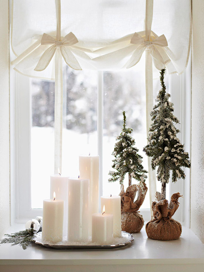 weihnachtsdeko auf einem kleinen kaffeetisch unter dem fenster weiße kerzen in verschiedenen größen vorhänge deko mit schleifen