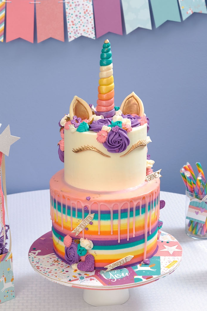 hier ist eine zweistöckige bunte einhorn torte mit einem weißen einhorn und mit einem regenbogenfarbenen horn - mit einer mähne aus lila, blauen und pinken rosen und mit einem lila herzen