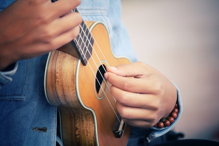 Mini Gitarre mit vier Saiten aus Naturfasern, Decke mit weißen Kanten, Musikspieler mit Denimhemd und Armbändern aus Holzperlen