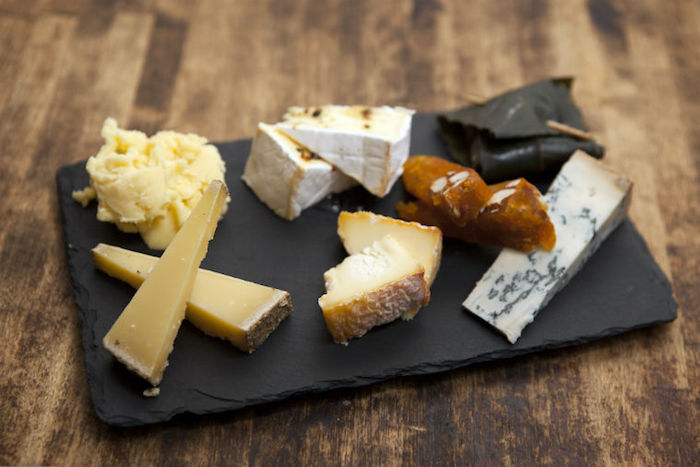schwarzer Servierteller aus Lavastein, gelegt auf einer Holzfläche, zwei Sorten harter Käse mit dicker Kruste, ein Stück Blaukäse mit Triangelform, Roulade aus Weinblatt mit einer Zahnstocher