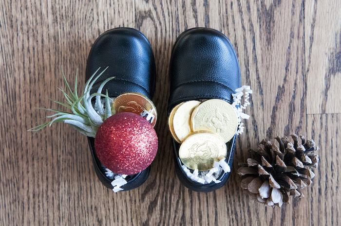 Nikolaus, schwarze Kinderschuhe aus Leder, gefüllt mit Schokomünzen und künstlichem Schnee, roter Weihnachtsbaumkugel mit Glitzer, Zapfen, Parkettboden