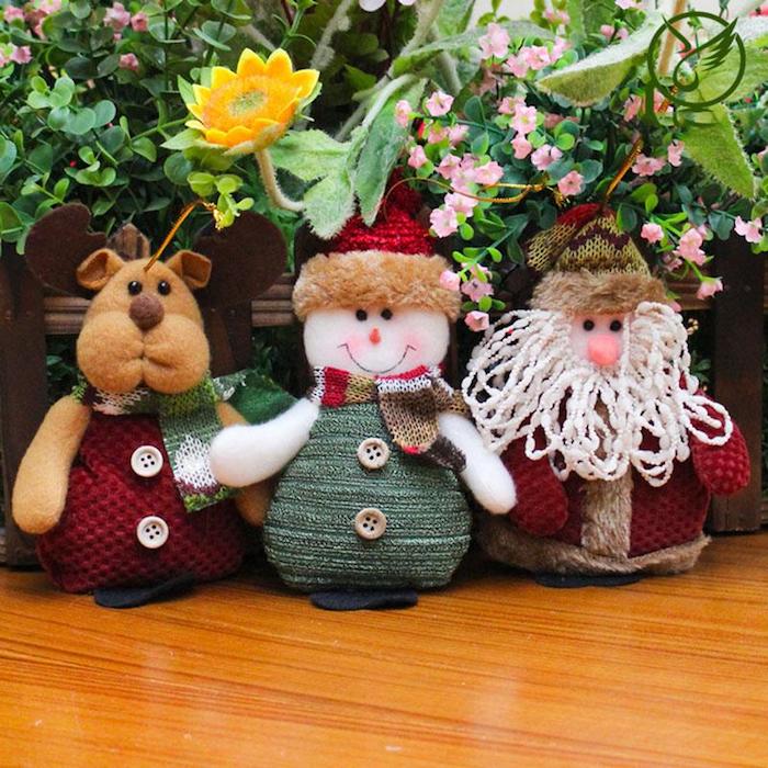 drei Strickfiguren - Rudolph mit einem roten Mantel mit weißen Knöpfen, ein lächelnder Schneemann mit rotem Winterhut, kariertem Schal und grünem Mantel mit grauen Knöpfen, der Weihnachtsmann mit roter Nase, künstliche Blumen, echte Pflanzen, Holztisch mit Lack