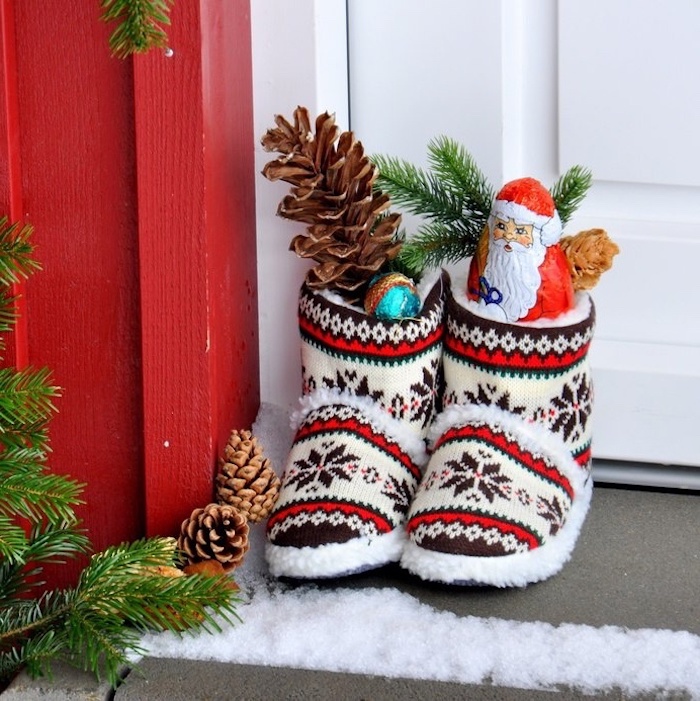Kinderschuhe für Zuhause mit Wintermotiven, gefüllt mit Zapfen und Schokoladenfiguren, weißer Holztür, roter Holzwand, Schnee vor der Eingangstür