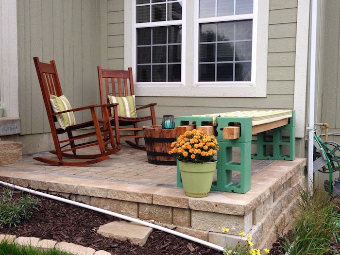 haus mit terrasse - zwei braune stühle mit grünen kissen - blumentopf mit orangen blumen - eine grüne gartenbank aus grünen pflanzsteinen 