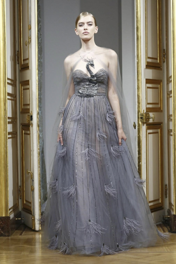 Yanina Couture-Kollektion - elegantes langes graues schulterfreies Kleid aus leichtem Stoff mit Applizierungen, Plissee-Oberteil mit Pfau-Motiv, getragen mit einem durchsichtigen Abendschal aus Tüll