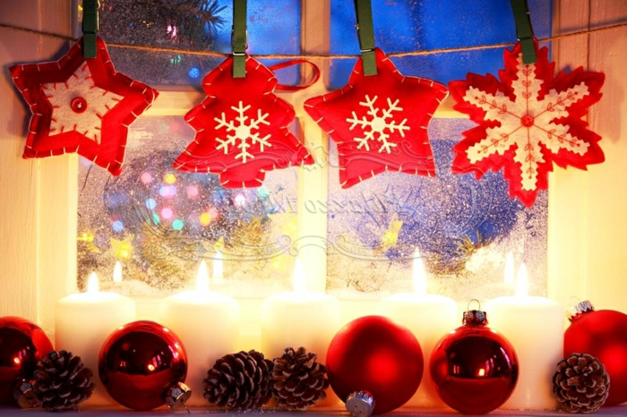 Schneeflocke aus weißer und roter Watte, verziert mit einer roten Glasperle in der Mitte, zwei Sterne in Weiß und Rot, kleiner Weihnachtsbaum aus roter Watte mit einer Schneeflocke aus weißer Watte, drei rote Christbaumkugeln mit Glanzüberzug, zwei rote Christbaumkugeln mit Mattüberzug, fünf brennende Kerzen aus weißem Kerzenwachs und vier Zapfen, rote weihnachtliche Dekoelemente, aufgehängt auf dem Fenster mit einem Bindfaden und grünen Wäscheklammern aus Holz