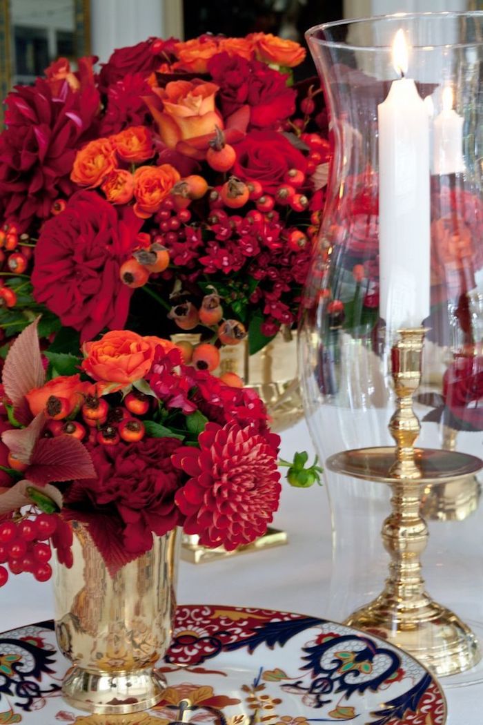 eine Kerze in hohem Glas, zwei Weihnachtsgestecke in goldenen Vasen voller rote Blumen