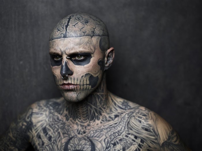 ein tätowierter unger mann mit vielen tattoos und mit einer tätowierung mit einem großen weißen totenkopf mit weißen zähnen und zwei schwarzen augen