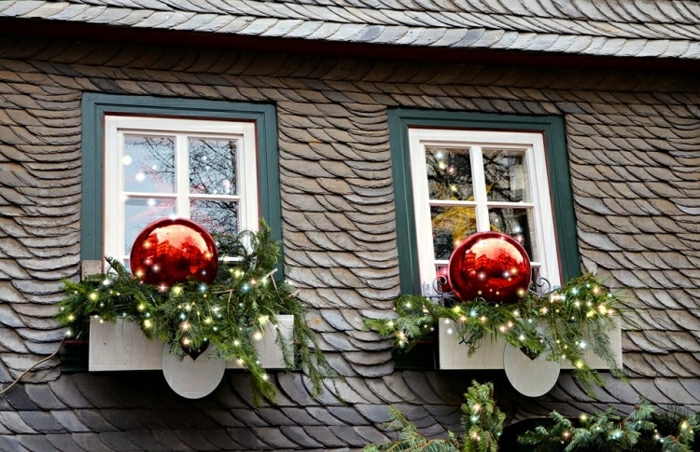 Holzhaus mit Schrägdach und zwei kleinen Fenstern mit Doppellrahmen, äußere Fenstersimse dekoriert mit Nadelbaumzweigen mit Lichterketten und zwei riesengroßen roten Weihnachtsbaumkugeln mit Glanz