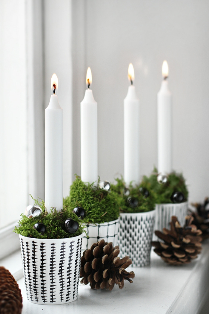 vier gezündete Kerzen aus weißem Wachs in vier schwarz-weißen Papierbechern, gefüllt mit frischem grünem Grass und kleinen schwarzen Kugeln