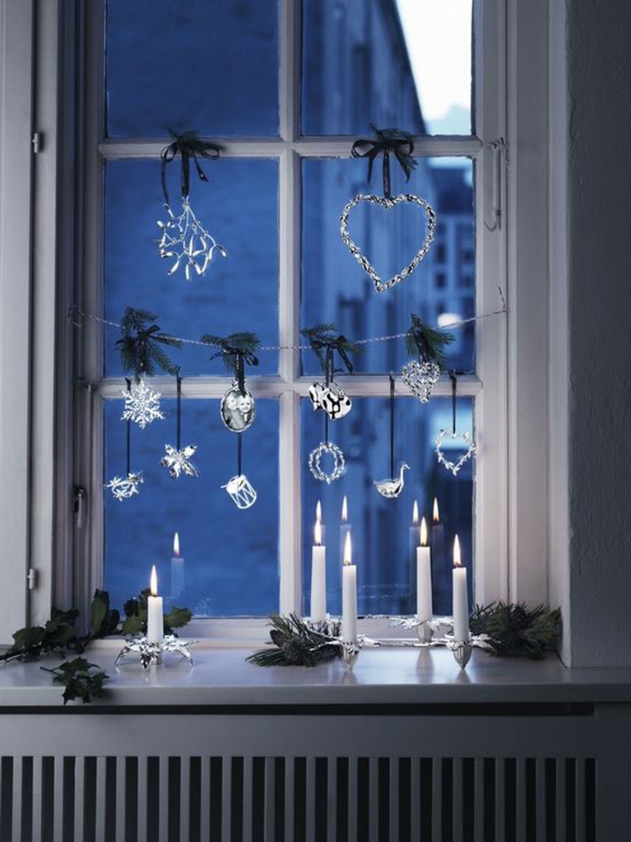 Fensterdeko für den Heiligen Abend - weiße Kerzen auf eleganten Kerzenhaltern, kleine Stücke Nadelbaumzweige, Fenster-Girlande aus Glasfiguren, Dekoelement in der Form eines Herzens mit kleinen LED-Lichtern und einem grünen Band, Heizungsverkleidung aus Holz