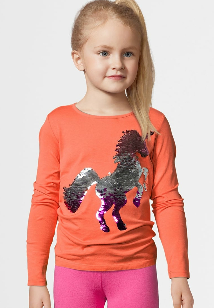 Süßes Kind mit langen blonden Haaren, Bluse in Apricot mit Einhorn-Applikation aus Pailletten