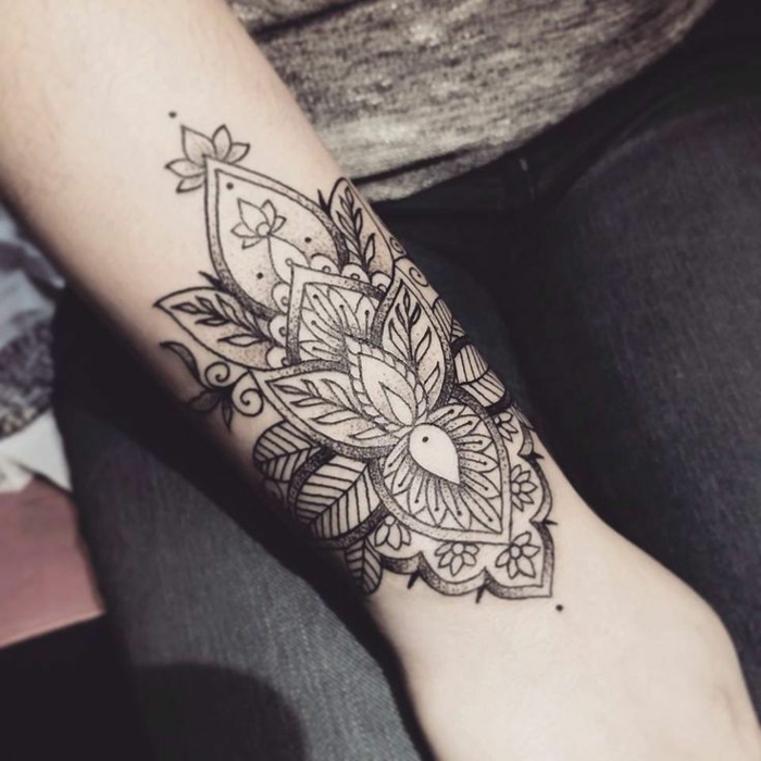 Handgelenk-Tattoo mit zwei kleinen grauen Lotusblumen, Spiralen und vielen Blättern, Verzierung mit kleinen Punkten, schwarze Jeans und graue Bluse