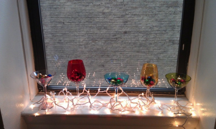 drei Cocktailgläser aus farbigem Glas, gefüllt mit kleinen Christbaumkugeln in verschiedenen Farben, zwei Weingläser aus rotem und gelbem Glas, Lichterkette mit stark brennenden Lichtern, Fenster mit dunklem Rahmen und Aussicht zum Nachbarhaus