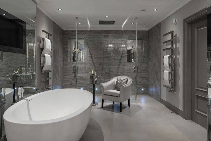elegante Badeinrichtung - Leder-Armstuhl in silberner Farbe mit einem grauen Deko-Kissen, Tuchaufwärmer an beiden Wänden, zwei Duschen mit dekorativen Nischen, Tür aus Massivholz