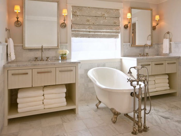 freistehende Badewanne mit vergoldeten Beinen mit Vintage-Motiven, Fensterrollos mit Blumenmotiv, Spiegel mit elegantem schmalen Rahmen, Haufen weiße Badtücher, weiße Blumen in einer Glasvase