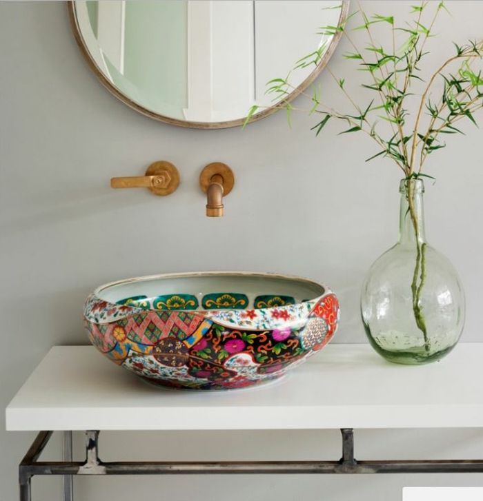 Spiegel in runder Form mit schmalem Edelstahl-Rahmen, dekorative Blumenvase aus grünlichem Glas mit einem frischen grünen Zweig, weißer Tisch mit Metallbeinen mit veraltetem Look