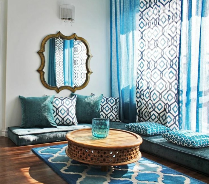 typische marokkanische Sitzecke in unterschiedlichen Blautönen und Weiß, weicher Musterteppich in drei Farben, großer Wandspiegel mit unregelmäßiger Form und ein vergoldetem Rahmen mit veraltetem Look, eine kleine Wandlampe darüber, niedriger runder Couchtisch aus Holz, blaue Glasvase mit runder Form, drei lange halbdurchsichtige Gardinen in Blau