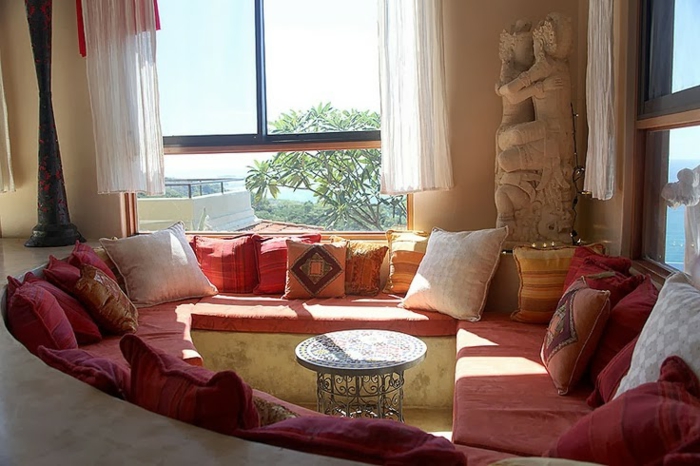Kaffee-Ecke im Wohnzimmer, Couch in Terrakotta-Farbe mit ovaler Form, runder Tisch aus Metall mit vielen Ornamenten, zwei große Fenster mit kurzen weißen Gardinen, Statue aus Stein in der rechten Ecke des Zimmers