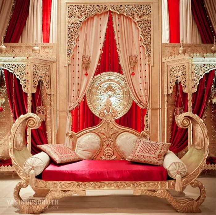 Einrichtung im Aladin-Stil - Liege mit Holzbeinen, gestrichen in Goldfarbe, Designer-Couch aus Massivholz, rundde Kissen mit Troddeln, Wandverkleidung mit Schnitzerei, runde Wanddeko aus Gold, lange rote schwerfallende Gardinen